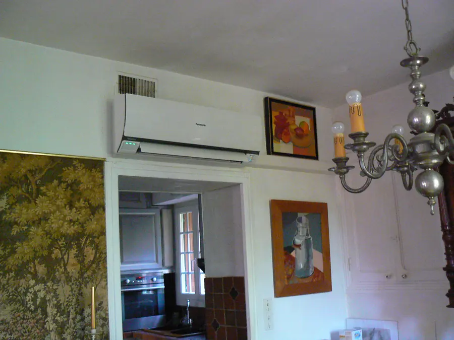 Unité intérieure murale posée au-dessus d'un passage, dans une salle à manger ancienne éclairée par un lustre.