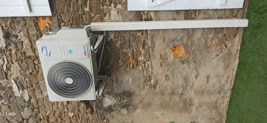 Unité extérieure trisplit Airwell fixée sur consoles contre un mur de pierre, la goulotte de liaison courant en façade.