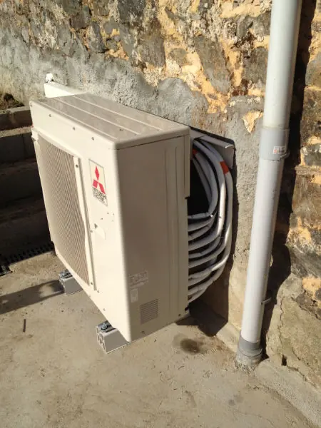Unité extérieure Mitsubishi Electric posée au pied d'un mur en pierre, liaisons frigorifiques lovées avant raccordement.
