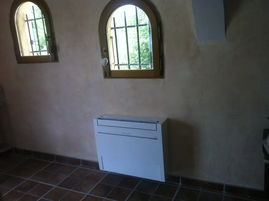 Console au sol Mitsubishi Electric posée sous deux fenêtres cintrées à barreaux, dans une pièce carrelée de tomettes.