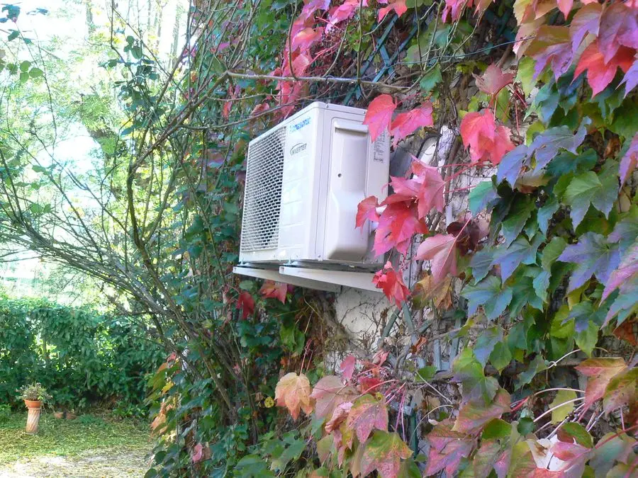 Unité extérieure de climatiseur fixée en façade, dissimulée par une vigne vierge aux feuilles rouges.